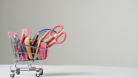 Close-up-of-miniature-shopping-trolley-school-materials-and-copy-space-on-grey-background