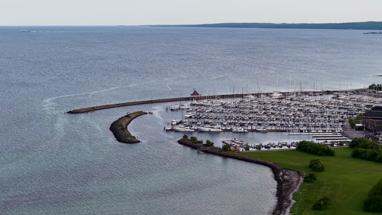 Droneoptagelse i luften af en lystbådehavn fyldt med sejlbåde ved siden af en lang bølgebryder, der strækker sig ud i det rolige hav