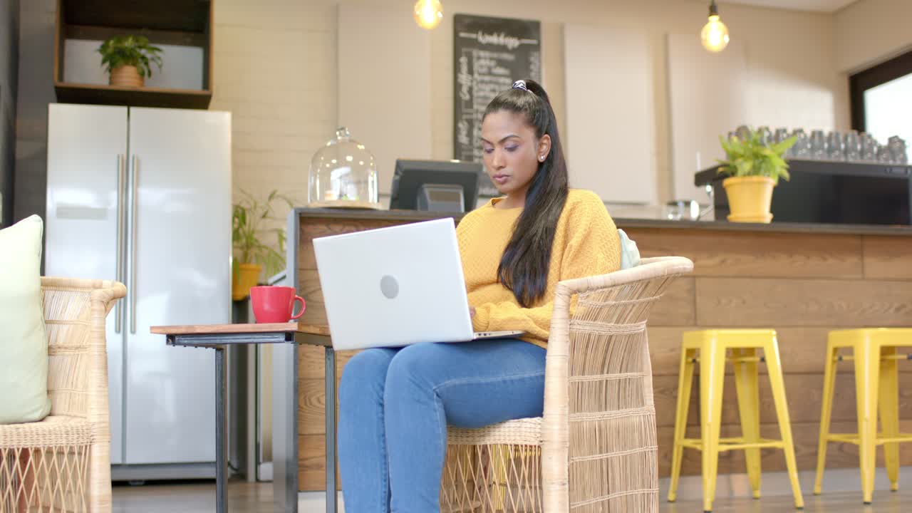 Indian woman typing on laptop at coffee shop with red mug, leaning and focusing on project