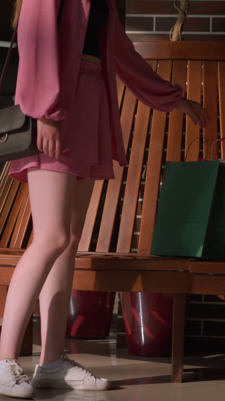 una dama con un traje rosa coloca sus bolsas de compras cerca de una silla de madera en un centro comercial, sostiene un bolso de mano negro y se sienta, con las piernas ligeramente cruzadas, con su mano izquierda descansando en el banco mientras se relaja