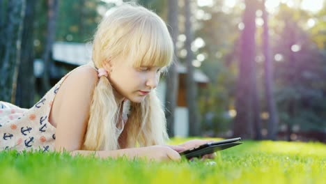 blonde girl  playing on the tablet, emotionally reacts. lying on the lawn in the backyard of the house. side view