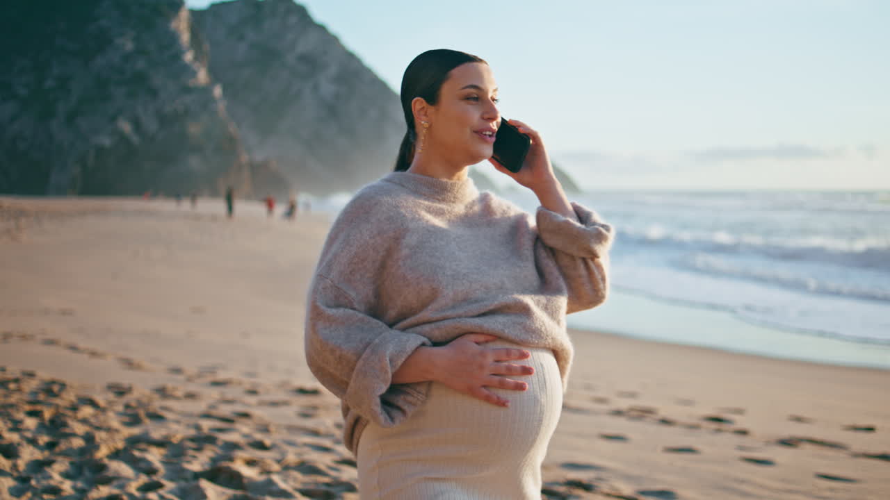 Pregnant girl calling beach caressing big belly. Future mother talking on phone.