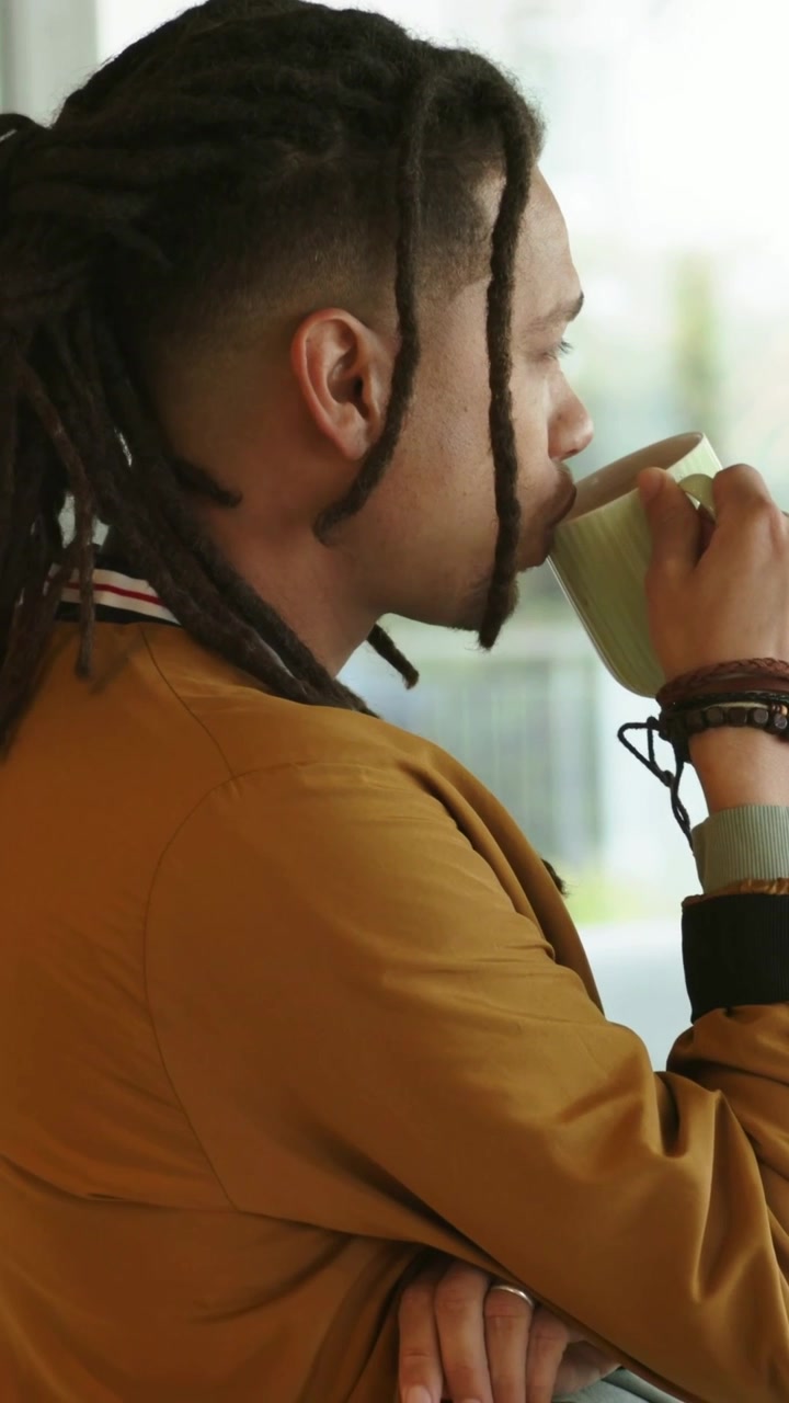 Vídeo vertical: Homem levantando caneca verde, bebendo e olhando pela janela, jaqueta mostarda, com espaço para texto.