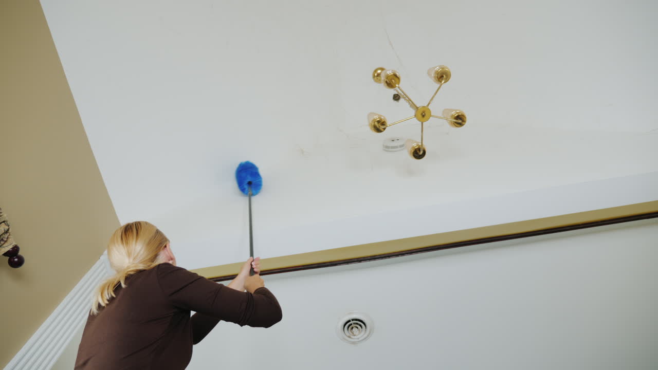 한 여자가 샹들리에 큰 집 청소 근처 천장에 있는 거미줄과 파편을 청소합니다