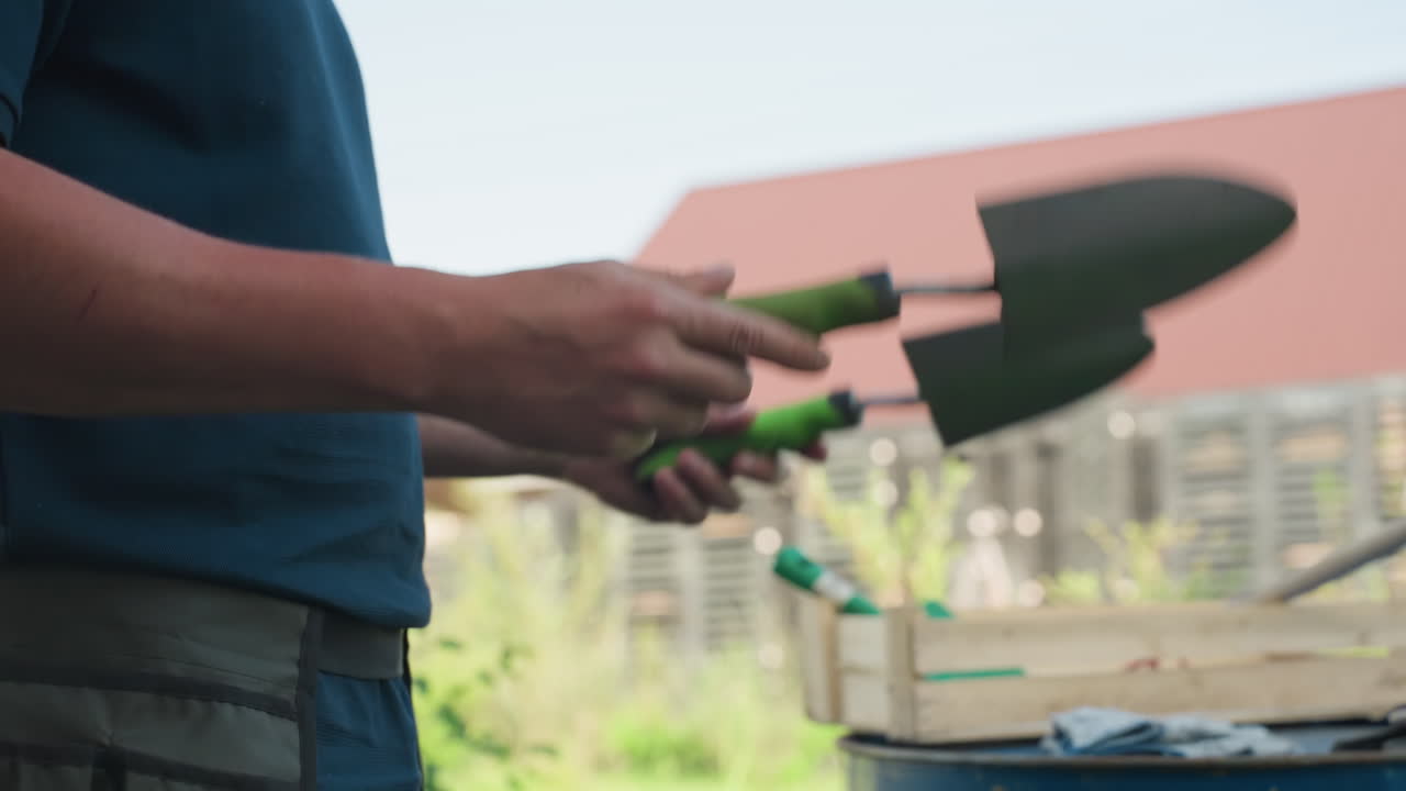 Primo piano di un contadino che sistema una paletta a mano, riponendo l'attrezzo nella cintura del grembiule in vita, con uno sfondo di giardino con una cassa di legno e un bidone blu nelle vicinanze, scena diurna primaverile, preparazione per la semina.