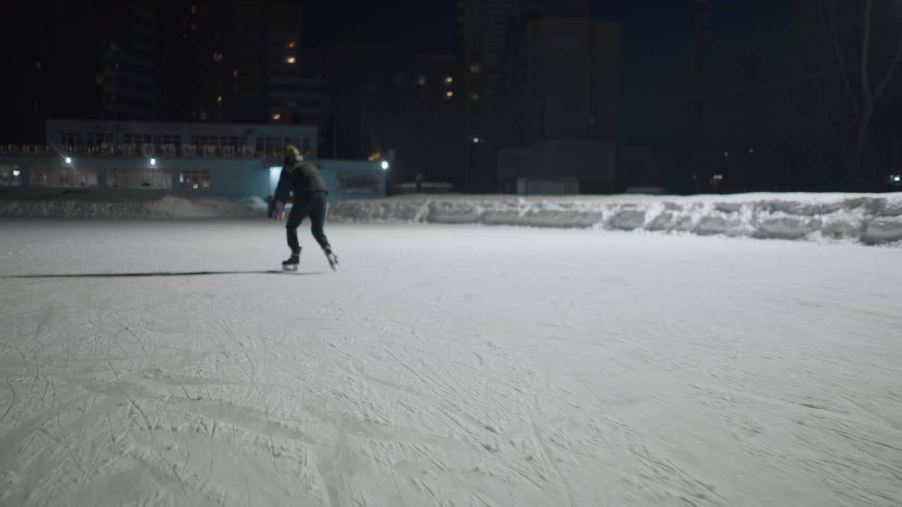 fast skater leans forward speeding across wide ice rink under bright overhead lights during night with urban buildings and snowbanks in softly lit background