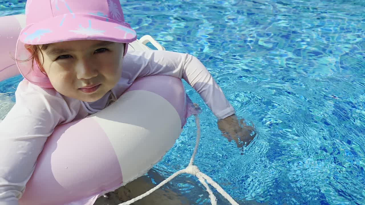 Close-up shot of a happy 5-year-old mixed Korean girl wearing a pink hat and floating on an inflatable ring, enjoying summer play in a bright blue swimming pool
