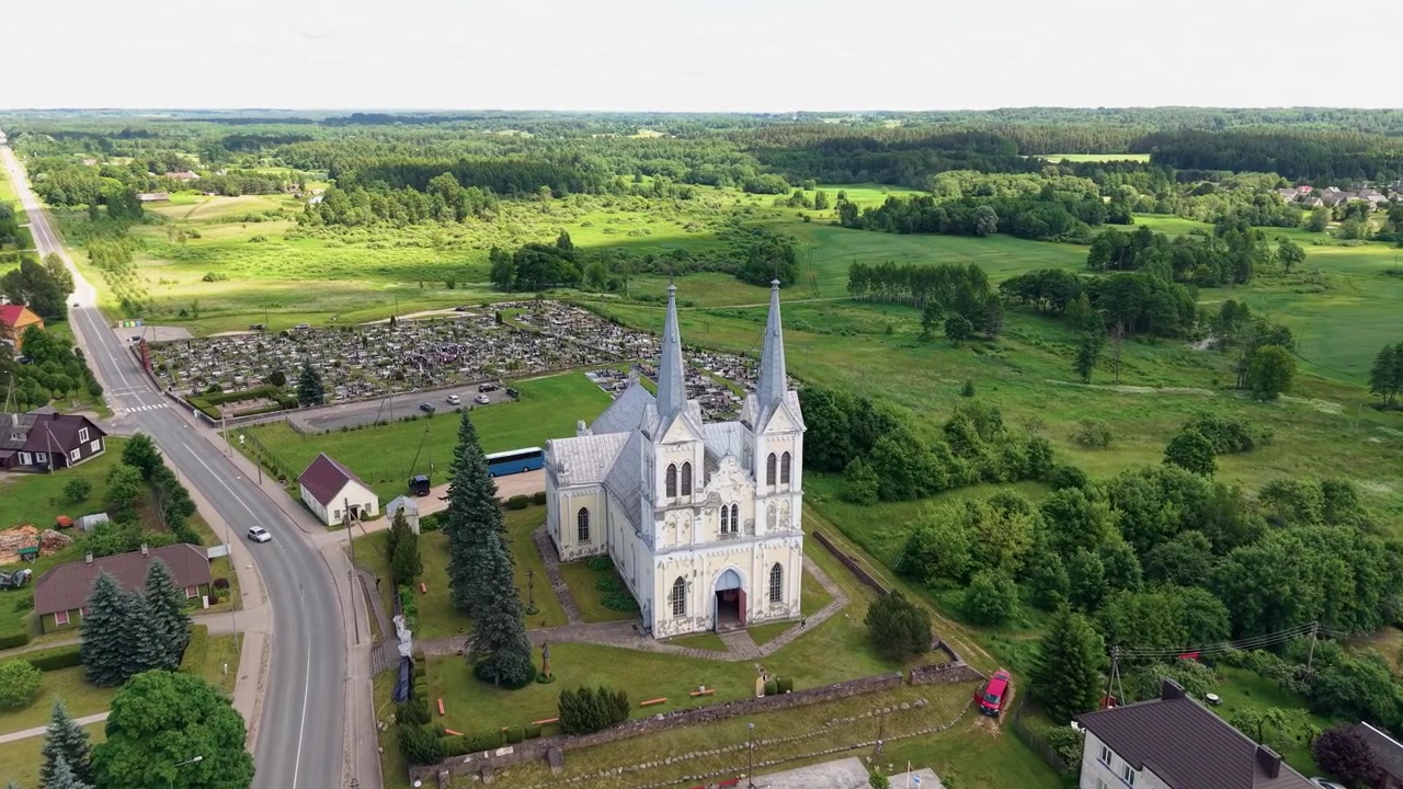 Drönarvideo visar Obeliai Sankta Annas kyrka med dubbla spiror, omgiven av gröna fält och landsvägar i mjukt dagsljus, vilket framhäver fridfull litauisk arkitektur och landskapets skönhet