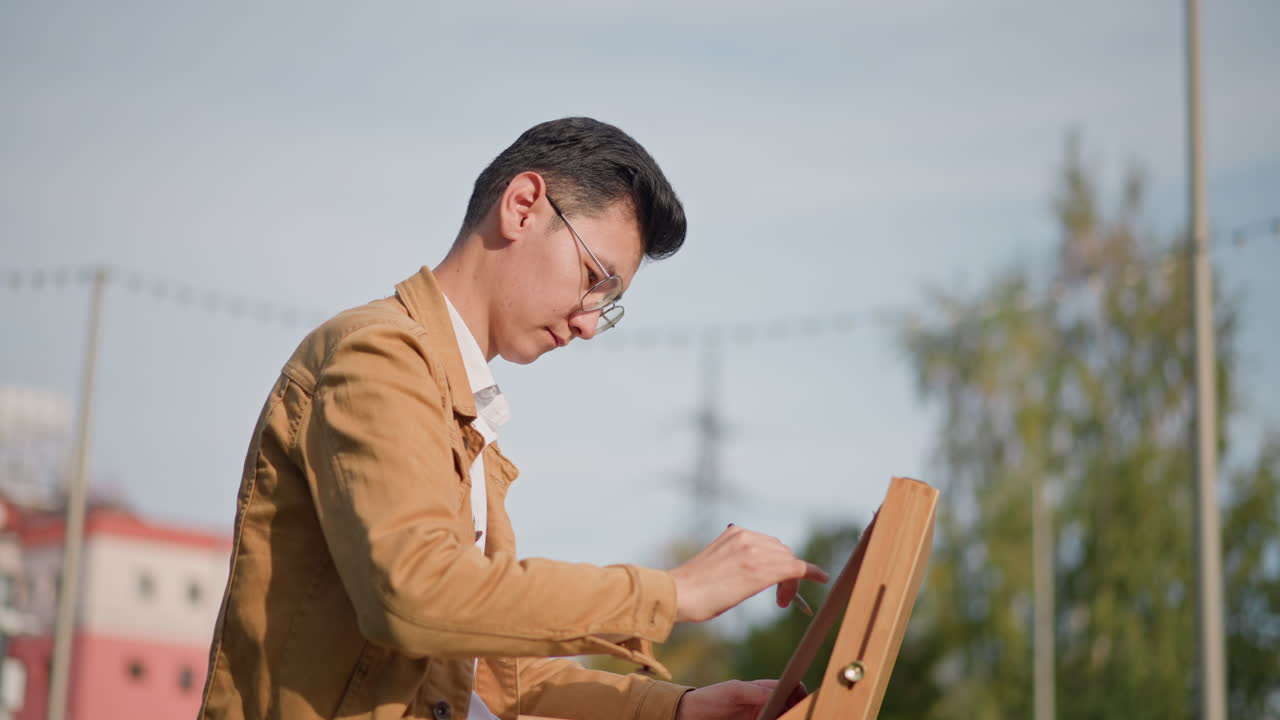 Aziatische schilder (man) buiten bij een ezel, zonnig park met bomen en een verre skyline van de stad, bruine jas en bril dragend terwijl hij een schets maakt op een canvas in een houten openluchtbox, geconcentreerde uitdrukking