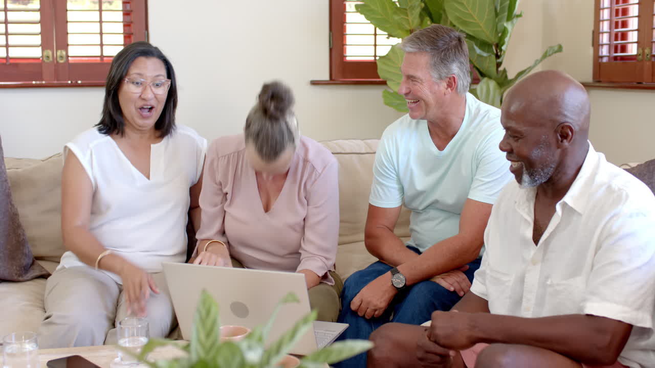 Diverse senior group laughing and chatting together on sofa with laptop, at home
