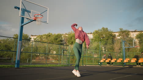 lady in outdoor court jumps, throws volleyball up, waits for it to land, then catches it, basketball hoop visible in background with people seated in the background