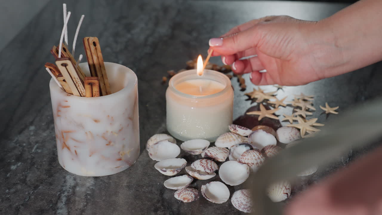 Hand view of young woman striking match to light decorative white candle on marble table, surrounded by scattered seashells, starfish, snail shells, and wick sticks