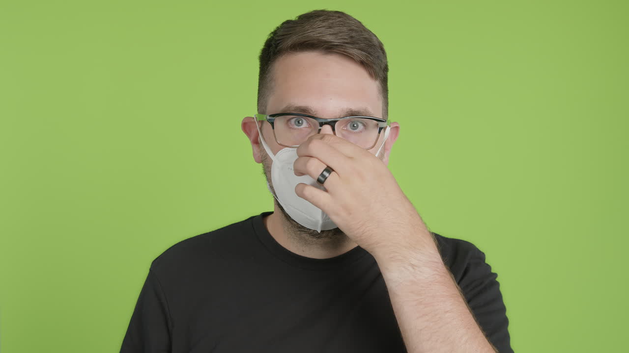 안경을 쓴 남자는 보호용 kn95 얼굴 마스크를 착용하고 녹색 화면 크로마 키에서 카메라를 들여다봅니다.