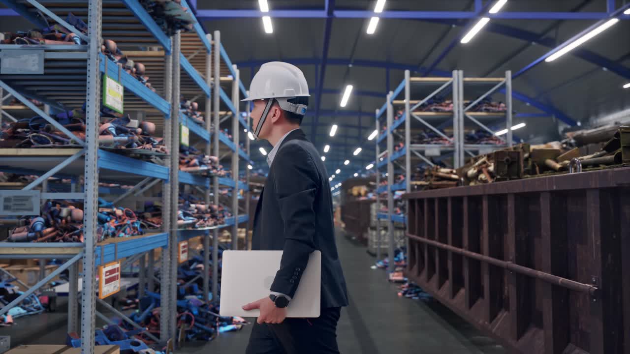 Vista laterale di un uomo d'affari asiatico che tiene un computer mentre cammina in un magazzino industriale di rottami metallici e riciclaggio.