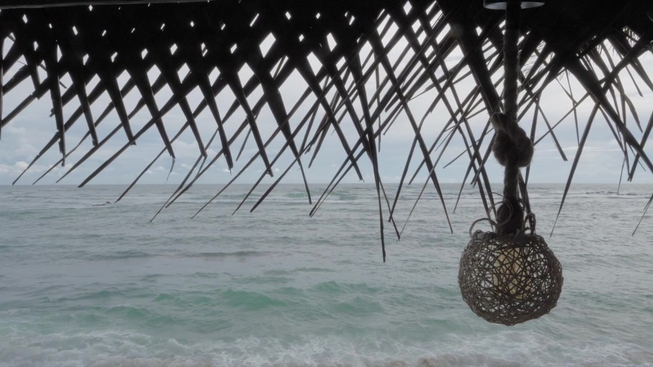 Vista da sotto una capanna sulla spiaggia con tetto in foglie di palma e una lampada a sospensione in vimini, che si affaccia sulle onde impetuose dell'oceano sulla costa tropicale di Mirissa, Sri Lanka