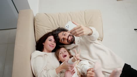 Young-family-are-making-a-selfie,-posing-while-lying-on-a-sofa-in-a-living-room,-high-angle-view