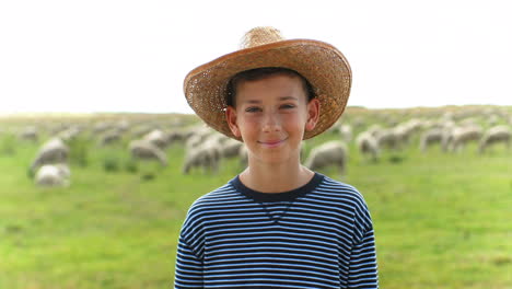 close-up view of caucasian teen boy in hat looking at camera in green field while the sheep flock is grazing