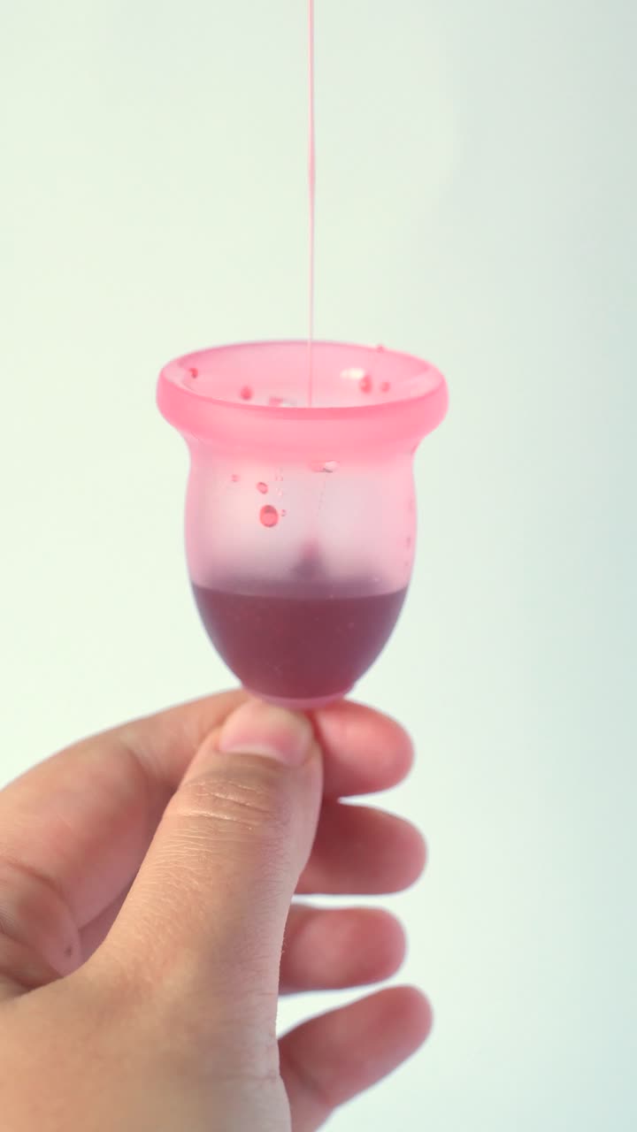 Donna che dimostra di versare liquido rosso in una coppetta mestruale rosa, simulando il flusso mestruale, isolata su sfondo bianco.