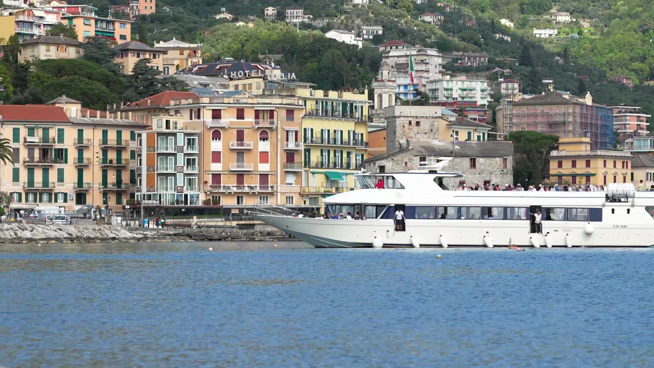 Scena vivace sul lungomare a Rapallo, Italia, con colorati edifici storici, il Castello di Rapallo e un moderno traghetto che scivola sull'acqua blu calma sotto una luce diurna soffusa. Urbano, vivace, cinematografico.