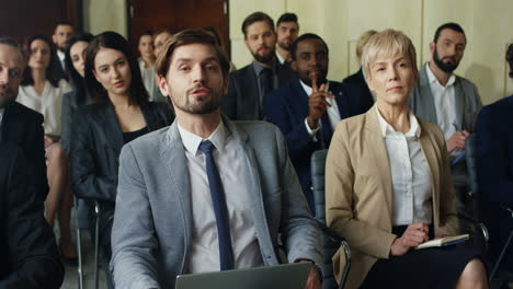 front view of caucasian businessman sitting among people in a conference room raising his hand talking to the camera