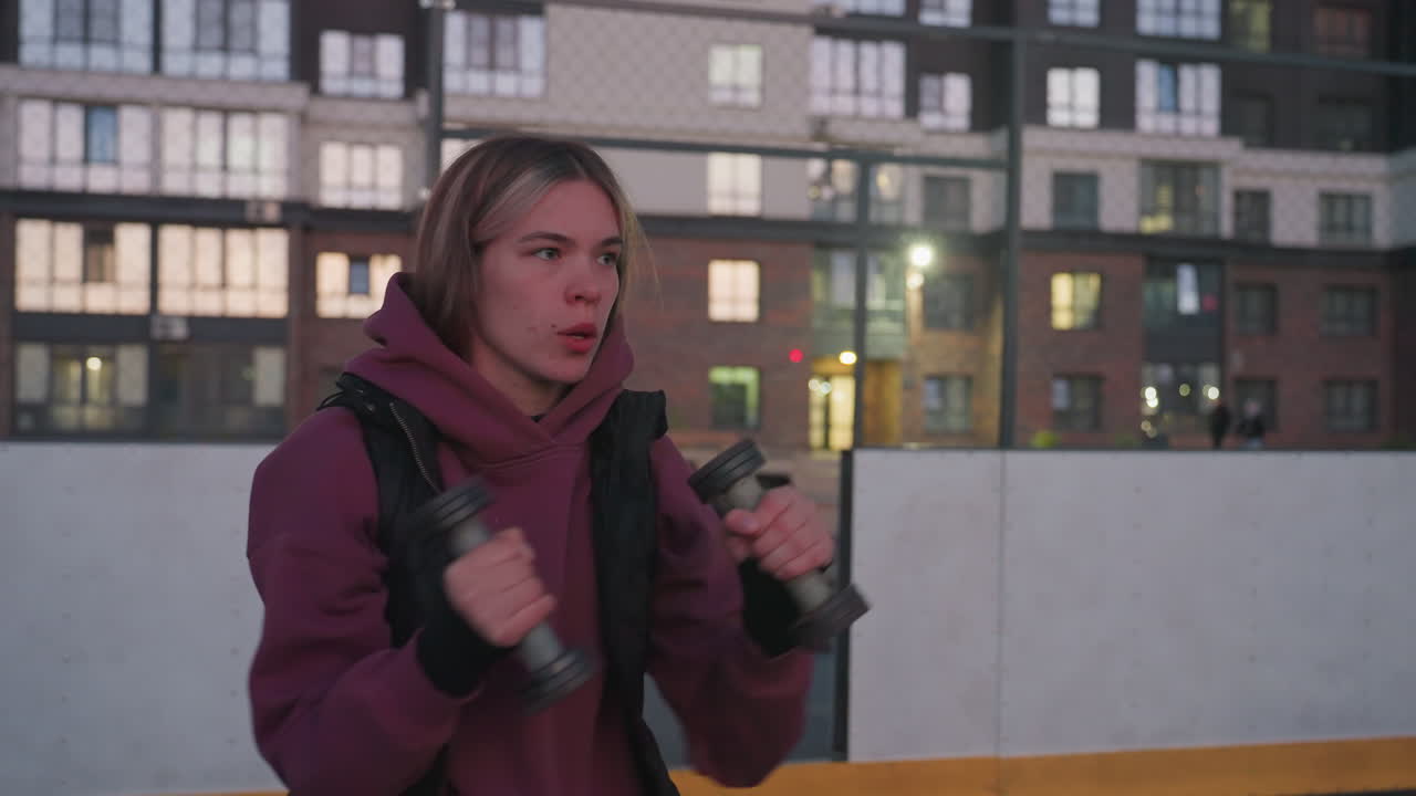 Exercise enthusiast stretching arms outward with dumbbells on black asphalt sports court beside white barrier with chain link fence against urban backdrop at dusk wearing maroon hoodie