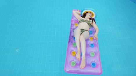 una joven con cabello oscuro en un colorido sombrero elegante tomando el sol en el colchón en la piscina