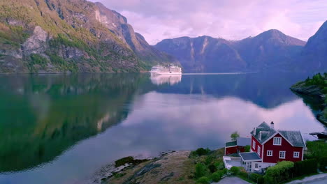 A-red-house-and-barn-with-a-view-of-the-fjord-and-reflective-water-surface-and-a-cruise-ship-in-Flam,-Norway