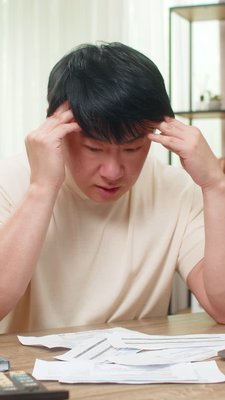 Asian man holding head over unpaid bills with calculator and laptop showing heavy financial stress