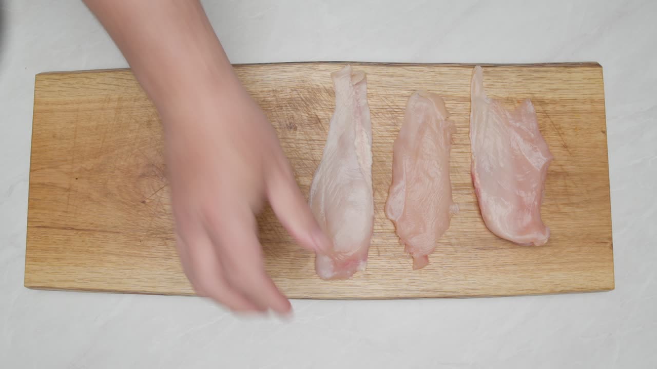 Primer plano cenital de una mano colocando filetes de pollo crudos sobre una tabla de cortar de madera