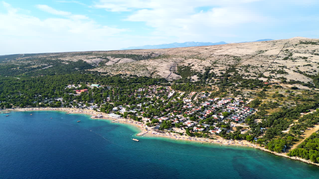 Turkosfärgat vatten möter den sandiga stranden vid Simuni strand, ön Pag, Kroatien, med klippiga kullar och berg i bakgrunden. Flyger från vänster till höger med drönare.