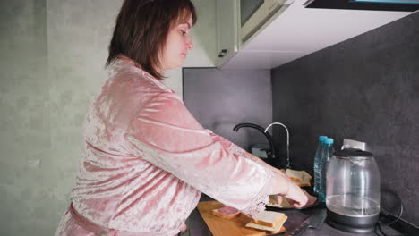 Caregiver in pink robe prepares sandwiches in modern kitchen, carefully placing slices of bread on cutting board next to plate with cheese and meat, water bottles, and electric kettle during morning routine