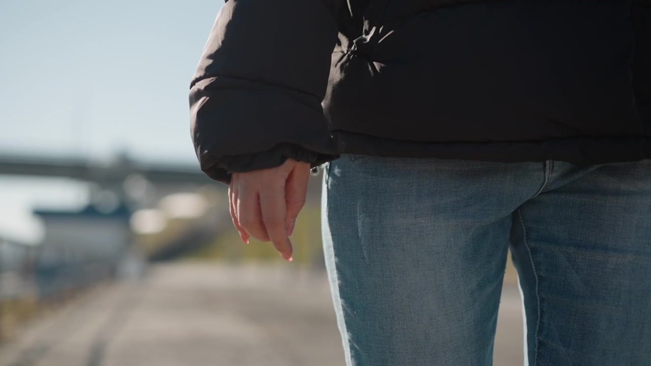 Una donna cammina all'aperto in un contesto informale, indossando jeans blu e un piumino nero. La telecamera segue la parte inferiore del suo corpo mentre si muove lungo un sentiero, con una luce naturale soffusa che illumina la scena.