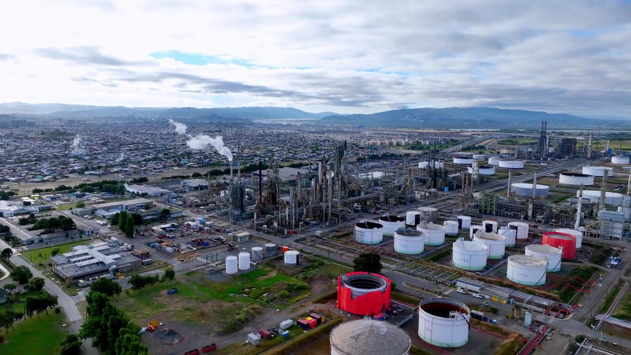 Toma aérea en retroceso de un complejo de refinería de petróleo que revela torres de destilación, columnas de vapor, tanques de almacenamiento y una densa zona residencial.