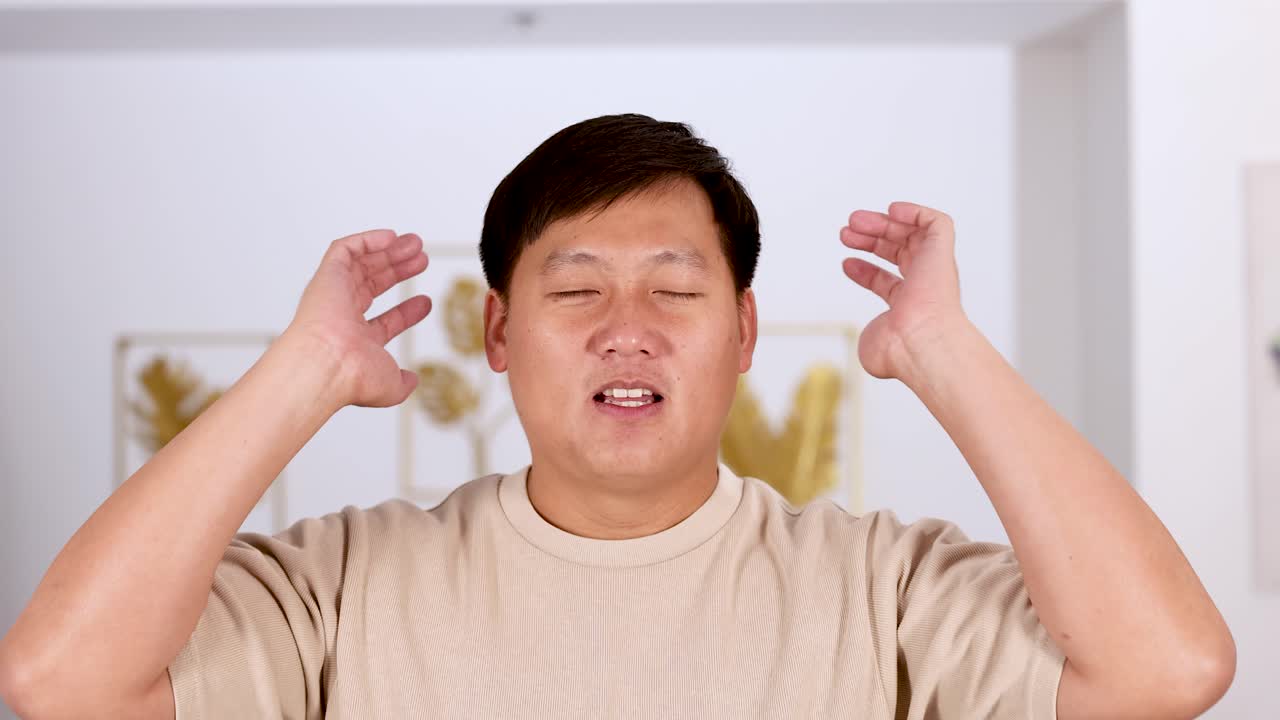 An Asian man covers his ears with both hands, expressing discomfort or frustration, in a brightly lit modern interior with soft, natural lighting and a static camera
