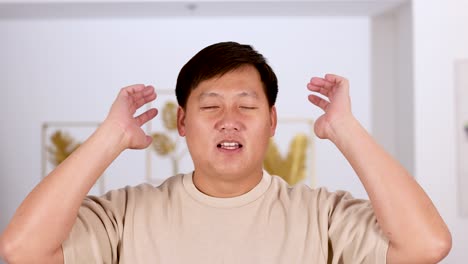 an asian man covers his ears with both hands, expressing discomfort or frustration, in a brightly lit modern interior with soft, natural lighting and a static camera