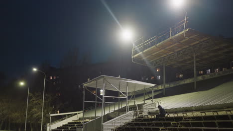 a man dressed in black and wearing a jacket sits alone in an empty stadium at night, with a soccer ball beside him with background illuminated by bright stadium light