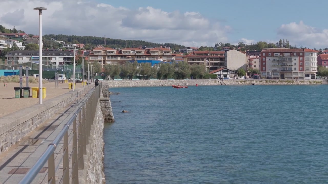 Baia di Plentzia Gorliz con una passeggiata in pietra, spiaggia sabbiosa, acqua blu calma ed edifici residenziali sotto un cielo luminoso