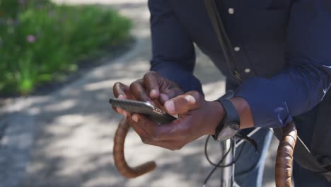 Afroamerikanischer-älterer-Mann-Mit-Gesichtsmaske-Und-Smartphone,-Während-Er-Sich-Auf-Seinem-Fahrrad-Auf-Der-Rechten-Seite-Lehnt