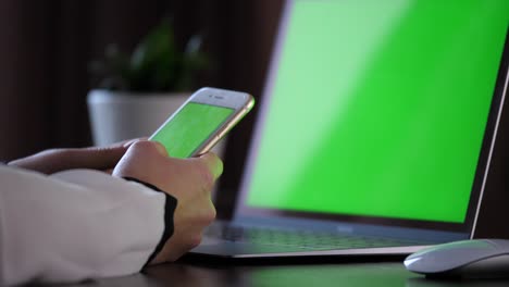 holding a smartphone with green screen, scrolling and then clicking with left finger on the upper left corner of the screen with a laptop with green screen in the back