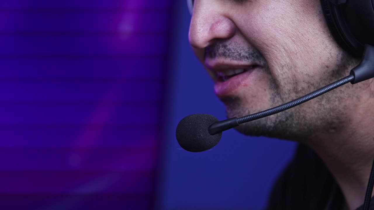 hombre hablando con un auricular mientras juega videojuegos mientras en el fondo ve una pantalla de televisión