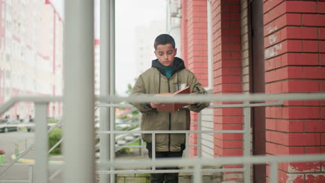 Young boy in hooded jacket studies while leaning on iron railing outside red brick building, with distant view of parked cars and residential blocks creating an urban learning atmosphere