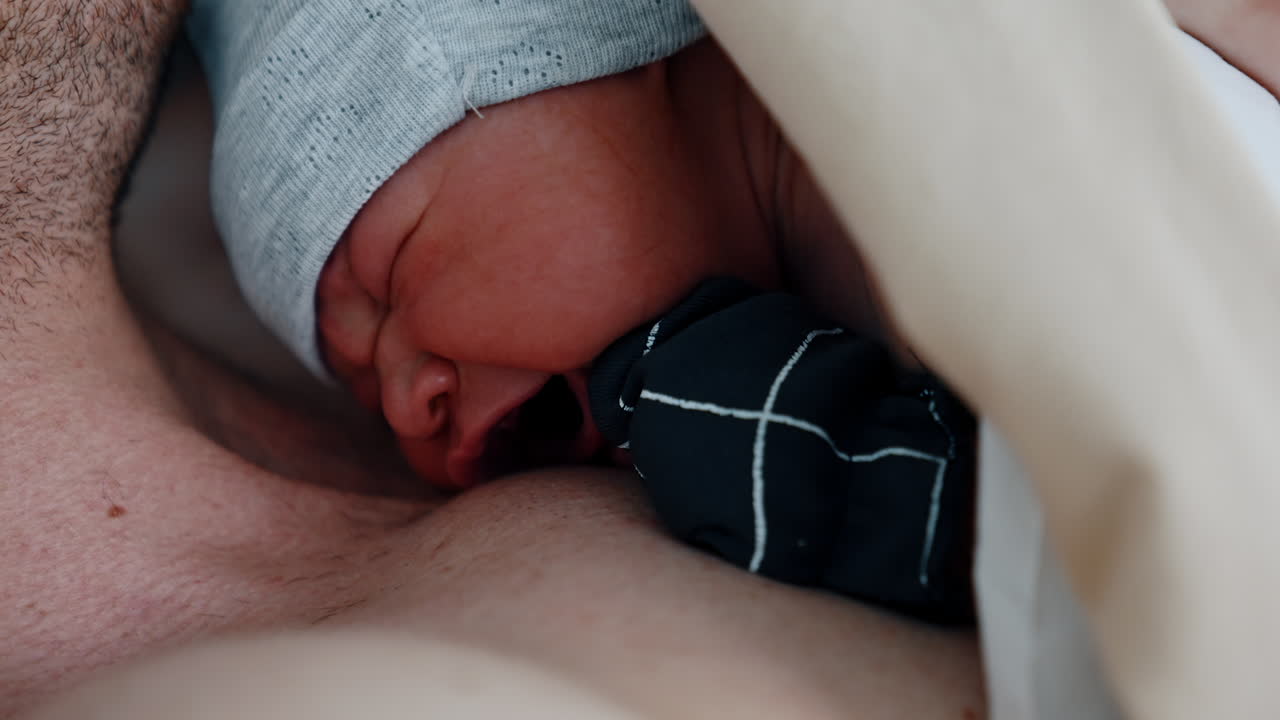 Tiny newborn is crying while lying on the chest of his dad. Baby opens eyes for the first time after being born. Close up.