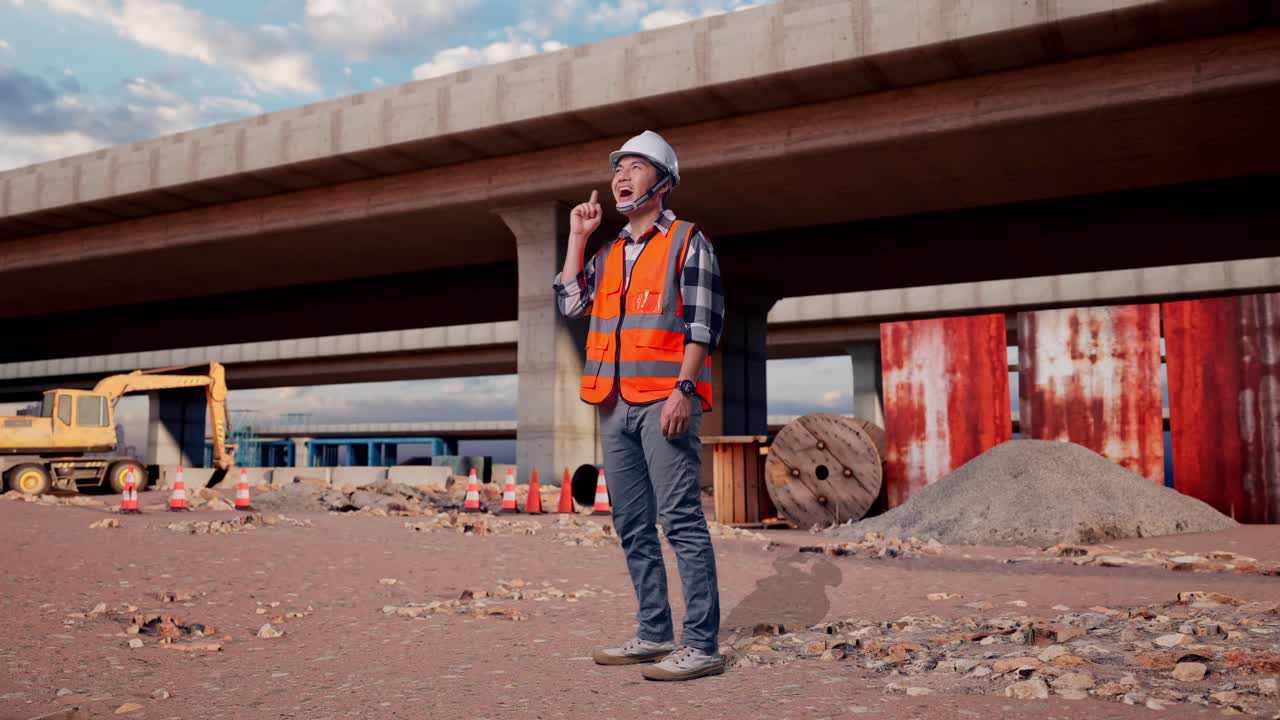 Full Body Side View Of Asian Male Engineer With Safety Helmet Thinking And Looking Around Then Raising His Index Finger While Standing At The Site of Highway Construction
