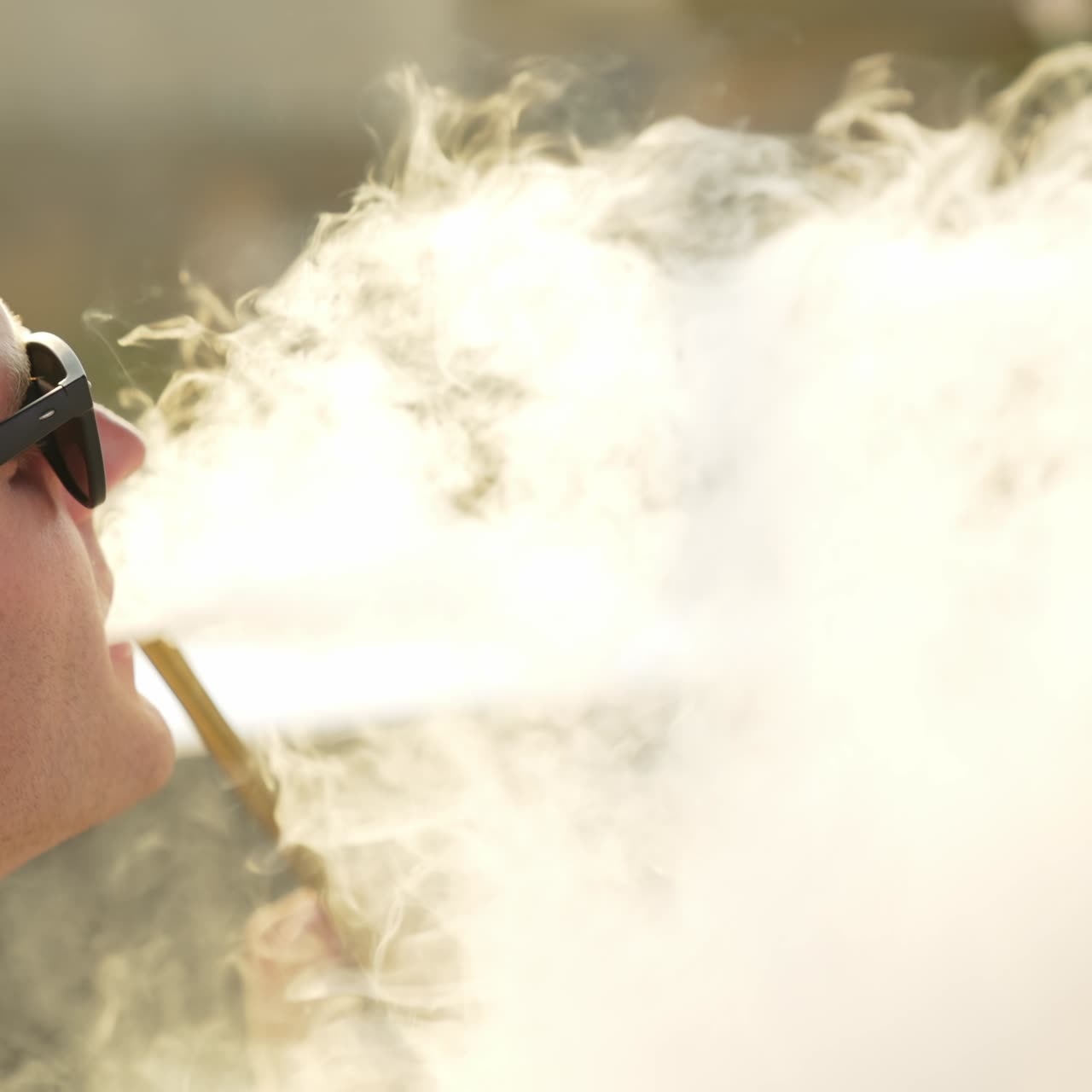 Caucasian young man breathing in smoke from pipe. Male in sunglasses makes a lot of thick white smoke from smoking hookah. Close up