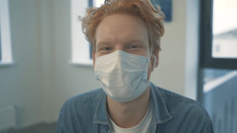 portrait of young handsome redhead male with face mask looking at camera and smiling indoor