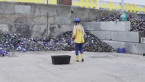 woman in hard hat with dreadlocks standing against the pile of broken glass, used bottles next to the wall. girl in yellow jacket throwing crashing old glass bottles for further recycling. slow motion