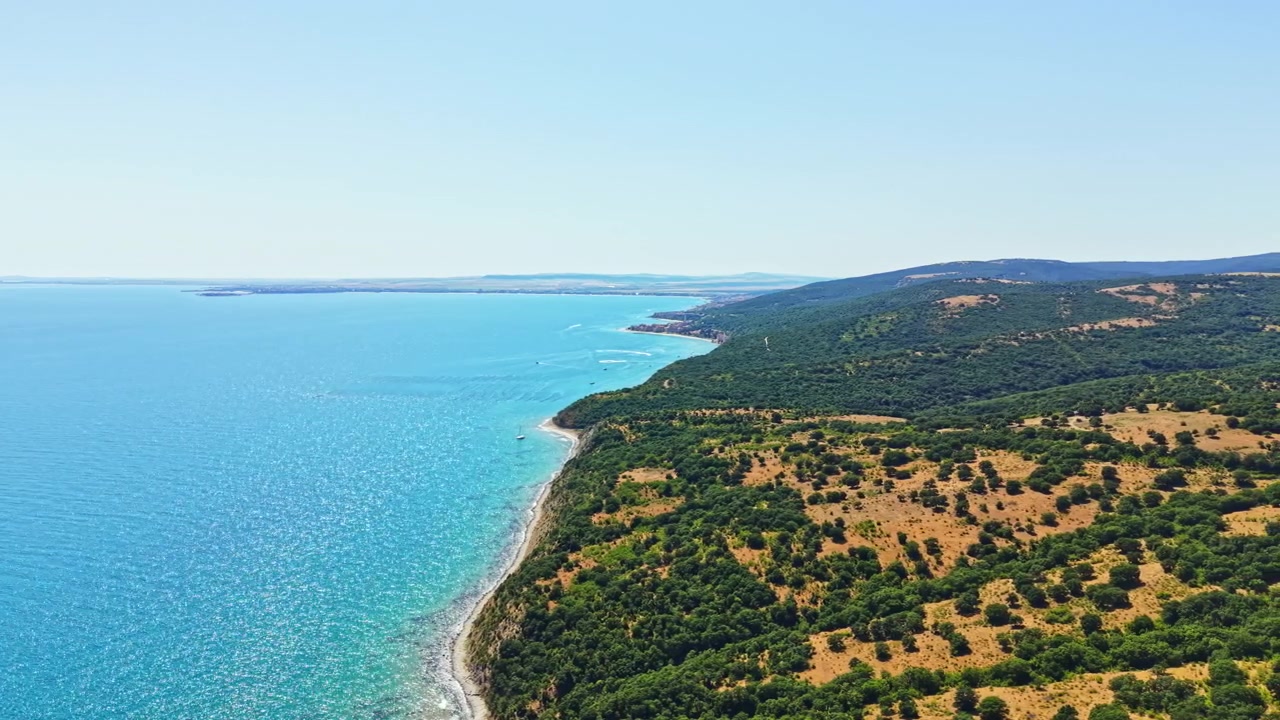 Veduta costiera con una collina verde e acque azzurre in una giornata di sole, sotto un cielo sereno e con onde leggere lungo la riva.