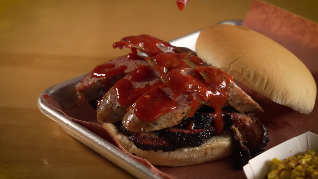 Bottiglia che versa salsa barbecue rossa su panino con salsiccia e brisket, ripresa ravvicinata in slow motion 4K.