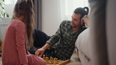 Side-view-of-a-brunette-man-with-stubble-in-a-checkered-shirt-playing-chess-with-his-little-daughter,-a-brunette-girl-in-a-pink-dress,-sitting-on-the-floor-and-leaning-on-a-gray-sofa-in-a-modern-apartment