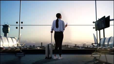 full body back view of asian woman with rolling suitcase in boarding lounge of airline hub, using smartphone while waiting for flight, airport terminal with airplane takes off outside the window